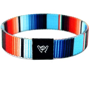 Könnte beinhalten: Ein kreisförmiges elastisches Armband mit einem lebendigen, mehrfarbigen Streifenmuster in leuchtendem Orange, Hellblau, Dunkelblau, Schwarz und Weiß. Ein kleines, rechteckiges schwarzes Stoffetikett ist angebracht und zeigt ein weißes Shaka-Handzeichen.