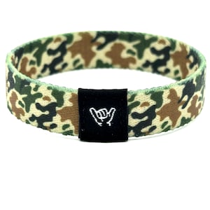 May include: A circular elastic wristband featuring a camouflage pattern in shades of green, brown, and tan. A small, rectangular black fabric tag is attached to the front, displaying a white embroidered shaka hand gesture icon.