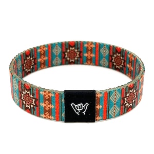 May include: A woven bracelet with a colorful geometric pattern in shades of orange, brown, blue, and white. The bracelet has a black adjustable buckle with a white "shaka" hand symbol.