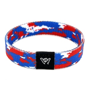 Può includere: Braccialetto in tessuto con motivo mimetico rosso, bianco e blu con una fibbia regolabile nera che presenta un gesto della mano "shaka" bianco.
