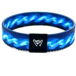 Op de afbeelding: Een blauwe elastische polsband met een herhalend patroon van lichtblauwe oceaangolven. Een klein, rechthoekig zwart stoffen label is in het midden van de band gestikt, met daarop een wit geborduurd icoon van een shaka-handgebaar.
