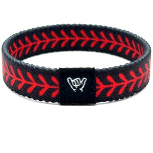 May include: A black fabric wristband featuring a continuous red stitched pattern resembling baseball laces. The band includes a small, rectangular black fabric tag centered on the front, displaying a white embroidered shaka hand gesture icon.