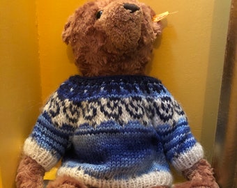 May include: A brown teddy bear wearing a blue and white knitted sweater with a traditional Scandinavian pattern.