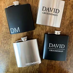Puede incluir: Cuatro matraces de acero inoxidable con texto grabado. Dos matraces son negros con texto blanco, uno dice "DM" y el otro dice "DAVID GROOMSMAN". Dos matraces son plateados con texto negro, uno dice "GROOMSMAN" y el otro dice "DAVID GROOMSMAN".