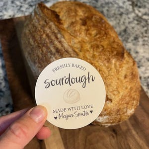 Puede incluir: Una hogaza de pan de masa madre recién horneado con una etiqueta redonda que dice "FRESHLY BAKED sourdough MADE WITH LOVE Megan Smith". El pan es de color marrón dorado con una corteza texturizada.