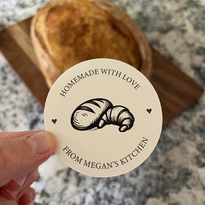 Puede incluir: Una pegatina redonda con el texto "Homemade with Love" y "From Megan's Kitchen" que enmarca una ilustraci&oacute;n de pan y croissant. Dos peque&ntilde;os corazones flanquean la ilustraci&oacute;n. Un pan se ve en el fondo.