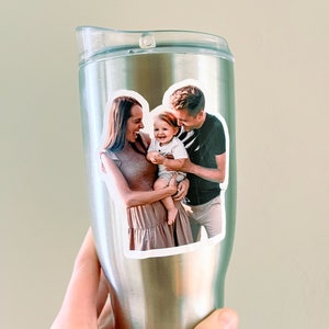 May include: A stainless steel travel mug with a photo sticker of a family of three. The photo shows a woman, a man, and a baby. The woman is wearing a brown dress and the man is wearing a black and white striped shirt. The baby is wearing a white shirt. The photo is in a white oval frame.