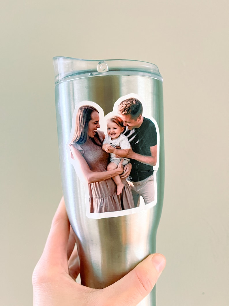 May include: A stainless steel travel mug with a photo sticker of a family of three. The photo shows a woman, a man, and a baby. The woman is wearing a brown dress and the man is wearing a black and white striped shirt. The baby is wearing a white shirt. The photo is in a white oval frame.