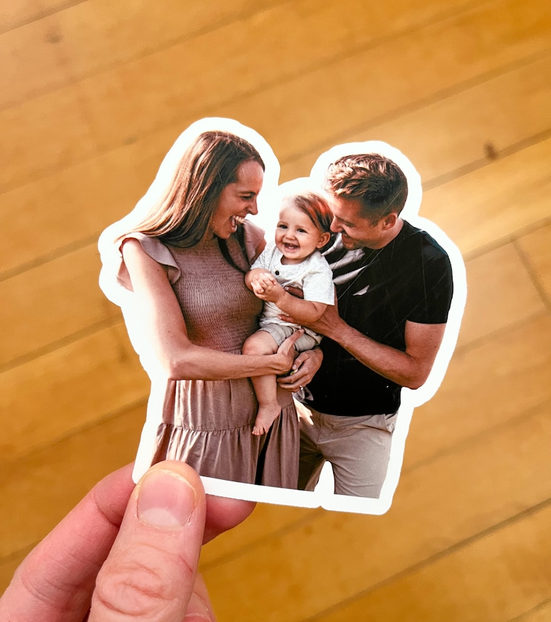 May include: A family portrait featuring a couple holding their baby. The woman is wearing a light brown dress and the man is wearing a black t-shirt and khaki pants. The baby is wearing a white shirt. The family is smiling and looking at each other.