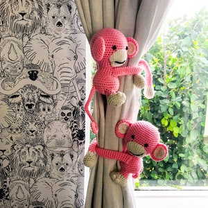 May include: Two pink crocheted monkey curtain tiebacks holding back a white curtain. The curtain is in front of a window with green foliage visible outside. The wall behind the curtain has a black and white animal print wallpaper.