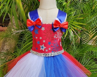 Op de afbeelding: Een rood, wit en blauwe tutu-jurk met een glinsterend lijfje met sterren en vuurwerk. De jurk heeft een blauwe satijnen strik op elke schouder en een zilveren paillettenband. Een bijpassend rood, wit en blauwe strik hoofdband is inbegrepen.