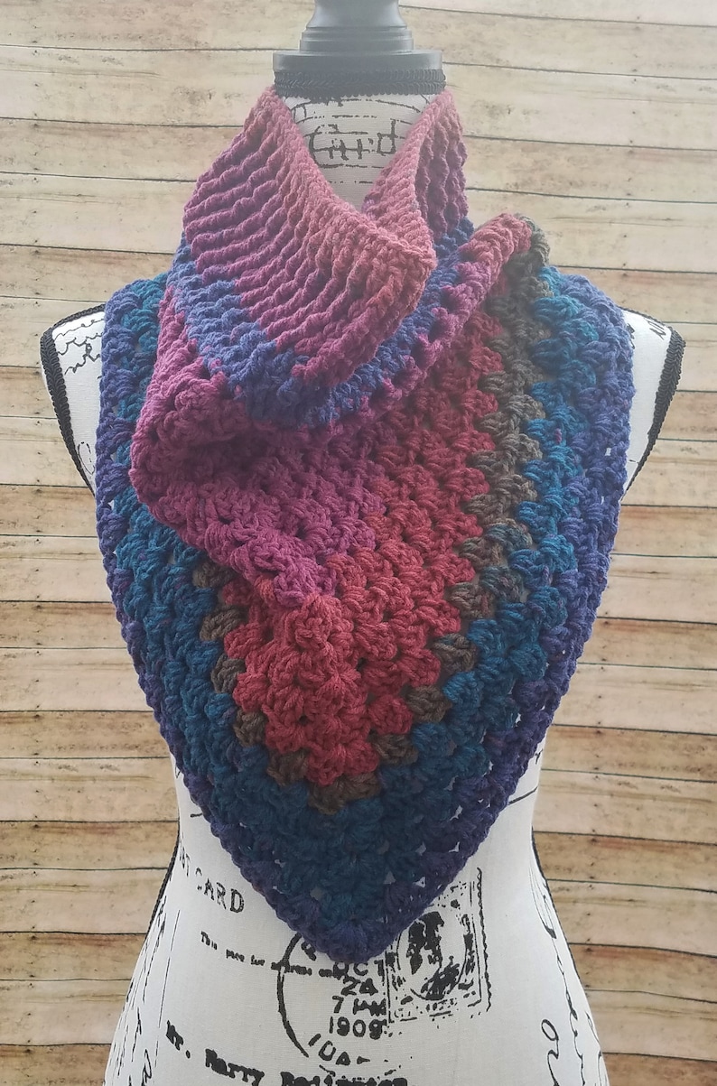 May include: A crocheted cowl in shades of purple, blue, and brown. The cowl is folded and draped over a white mannequin.