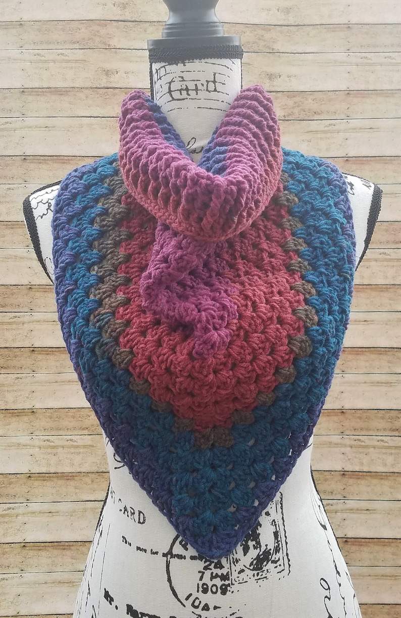 May include: A crocheted triangular scarf with a multi-colored design. The scarf is made with a variety of shades of blue, purple, and red yarn. The scarf is displayed on a white mannequin.