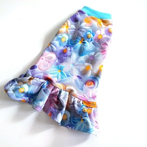 Puede incluir: Una camiseta para perro azul claro con estampado floral con flores rosas, moradas y amarillas. La camiseta tiene un dobladillo con volantes y un cuello turquesa.