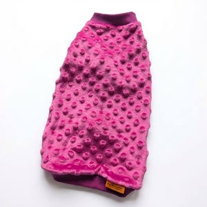 May include: A vibrant fuchsia dog sweater with a textured, dotted pattern. The sweater has a ribbed collar and hem in a darker shade of fuchsia. A small, rectangular tag with text is sewn on the bottom hem.