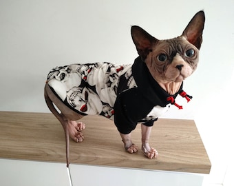 May include: A hairless cat wearing a black and white hoodie with a skull print. The hoodie has a red drawstring. The cat is standing on a wooden surface.