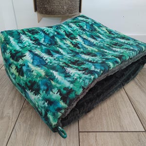 May include: A pet bed with a forest-themed pattern. The top layer features a vibrant green and blue tree design, while the interior is a dark gray, plush material. A small loop is attached to the corner.