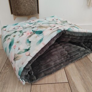 May include: A pet bed with a floral pattern on a light blue background. The interior is a soft, gray, ribbed fabric. The bed is open, inviting a pet to snuggle inside. The floral design includes pink blossoms and green leaves.