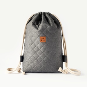 May include: A gray quilted drawstring bag with a brown leather patch that says "M" on it. The bag has white drawstrings.