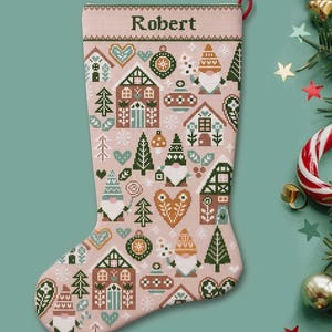 Op de afbeelding: Een lichtroze kerstkous met een kruissteekpatroon van kabouters, peperkoekhuisjes, hartjes en ornamenten. De naam Robert is bovenaan in donkergroene tekst geborduurd. Aan de zijkant zit een klein rood lusje om de kous op te hangen.