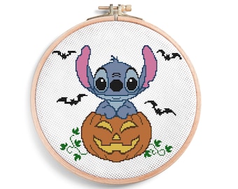 May include: A circular wooden embroidery hoop containing a cross-stitch pattern of a blue alien character sitting on top of an orange jack-o'-lantern. The design includes four small black bats flying in the background and green vine accents.