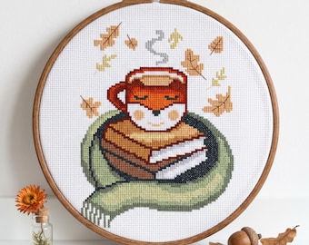 May include: A cross-stitch embroidery in a round wooden hoop featuring a fox-shaped mug resting on a stack of books, wrapped in a green scarf. The design includes falling autumn leaves in shades of orange and yellow, all stitched on white fabric.