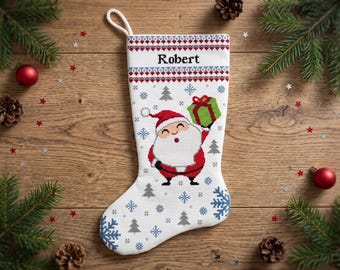 May include: A white cross-stitch Christmas stocking featuring a central Santa Claus figure holding a green gift box. The stocking has a red and blue geometric border at the top with the name Robert printed in black text and blue snowflakes.
