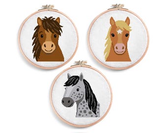 May include: Three cross-stitch embroidery hoops showing horse faces. One horse has a brown mane, another has a blonde mane with a white star, and the third is grey with a black mane. The hoops are set against a white background.