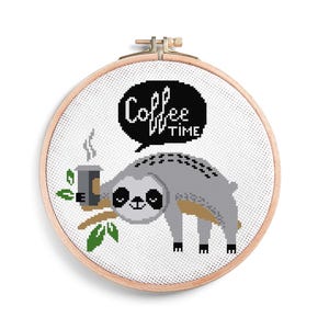 Puede incluir: Bordado de punto de cruz en un aro de madera que muestra un perezoso gris sosteniendo una taza de café en una rama. El perezoso sonríe, y una burbuja de diálogo dice "Coffee Time" en blanco y negro.