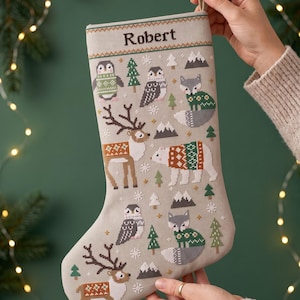 Puede incluir: Un calcetín navideño de color gris claro con un patrón de punto de cruz de varios animales de invierno, incluyendo renos, un oso polar, búhos y zorros con suéteres. El nombre Robert está bordado en texto marrón oscuro en la parte superior.