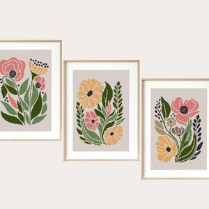 May include: Three framed floral art prints. Each print features a stylized floral arrangement in shades of pink, yellow, and green against a neutral background. The frames are a light wood color.