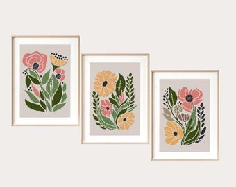 Könnte beinhalten: Drei gerahmte Kunstdrucke mit Blumenmotiven. Jeder Druck zeigt ein stilisiertes Blumenarrangement in Rosa-, Gelb- und Grüntönen vor einem neutralen Hintergrund. Die Rahmen sind hellholzfarben.