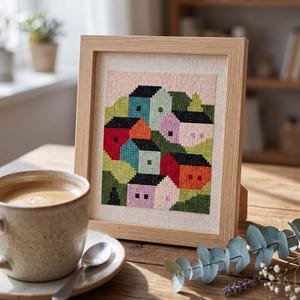 May include: A framed cross-stitch embroidery featuring a stylized village scene with colorful houses in shades of red, orange, blue, and purple, set against a light pink sky and green hills, all enclosed in a light-toned wooden frame.