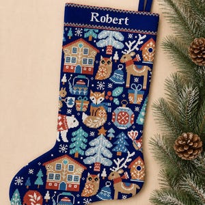 Puede incluir: Un calcetín navideño azul oscuro con un patrón de punto de cruz de casas, renos, zorros, búhos, pinos y regalos. El nombre Robert está bordado en blanco en la parte superior. El calcetín incluye un lazo azul oscuro para colgarlo.