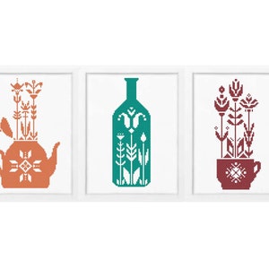 May include: Three framed cross-stitch patterns featuring stylized floral designs in a teapot, bottle, and cup. The teapot is orange, the bottle is teal, and the cup is maroon.