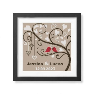 Op de afbeelding: In een zwarte lijst geplaatst kruissteekkunstwerk met twee rode vogels op een bruine tak, omringd door witte harten. De namen "Jessica & Lucas" en de datum "12.09.2023" zijn zichtbaar.
