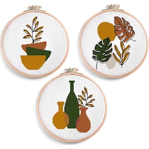 Puede incluir: Tres obras de arte en punto de cruz enmarcadas con diseños botánicos. Una presenta cuencos apilados con una planta, otra hojas y un círculo, y la última jarrones y follaje. Los marcos son redondos y de madera.