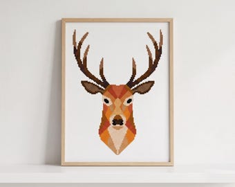 May include: A cross-stitch pattern of a deer head featuring geometric shapes in shades of orange, brown, and tan. The deer has large, dark brown antlers and is centered on a white grid background, enclosed within a light-colored wooden frame.