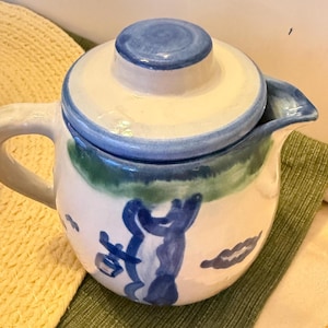 May include: A ceramic teapot with a lid, painted with blue, green, and white colors. The teapot has a handle and a spout. The design includes blue abstract shapes and a green band around the middle. The lid is blue.