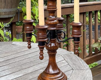 May include: A wooden candelabra featuring a dark brown polished finish with black metal accents. The piece has a central pillar and four curved arms, each holding a tall, slender, light beige taper candle. The base is wide and circular.