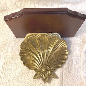 May include: A decorative wall shelf with a dark brown wooden top and a gold-colored, seashell-shaped brass accent piece. The shelf has a classic design, suitable for displaying small items or adding a touch of coastal decor.