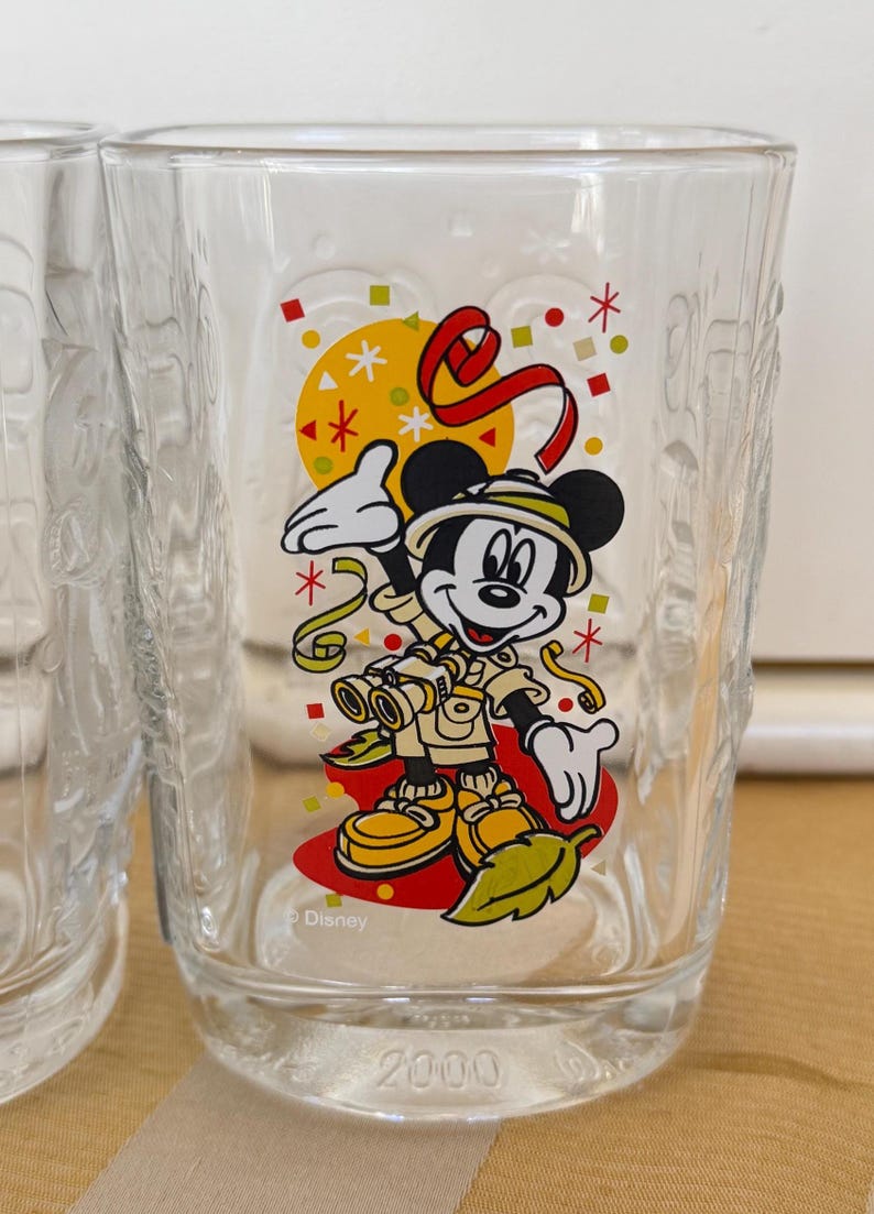 以下が含まれることがあります： サファリ衣装のミッキーマウスが双眼鏡を持っているカラフルなグラフィックが特徴の透明なガラス。デザインには、黄色の太陽、赤いリボン、紙吹雪が含まれています。ガラスの底には「2000」とディズニーのロゴが刻印されています。