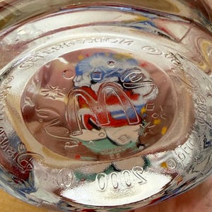 以下が含まれることがあります： 丸みを帯びた底を持つ透明なガラス瓶。底には、赤と黄色のマクドナルドのロゴがあり、カラフルな画像で囲まれています。「5000」の数字が底にエンボス加工されています。