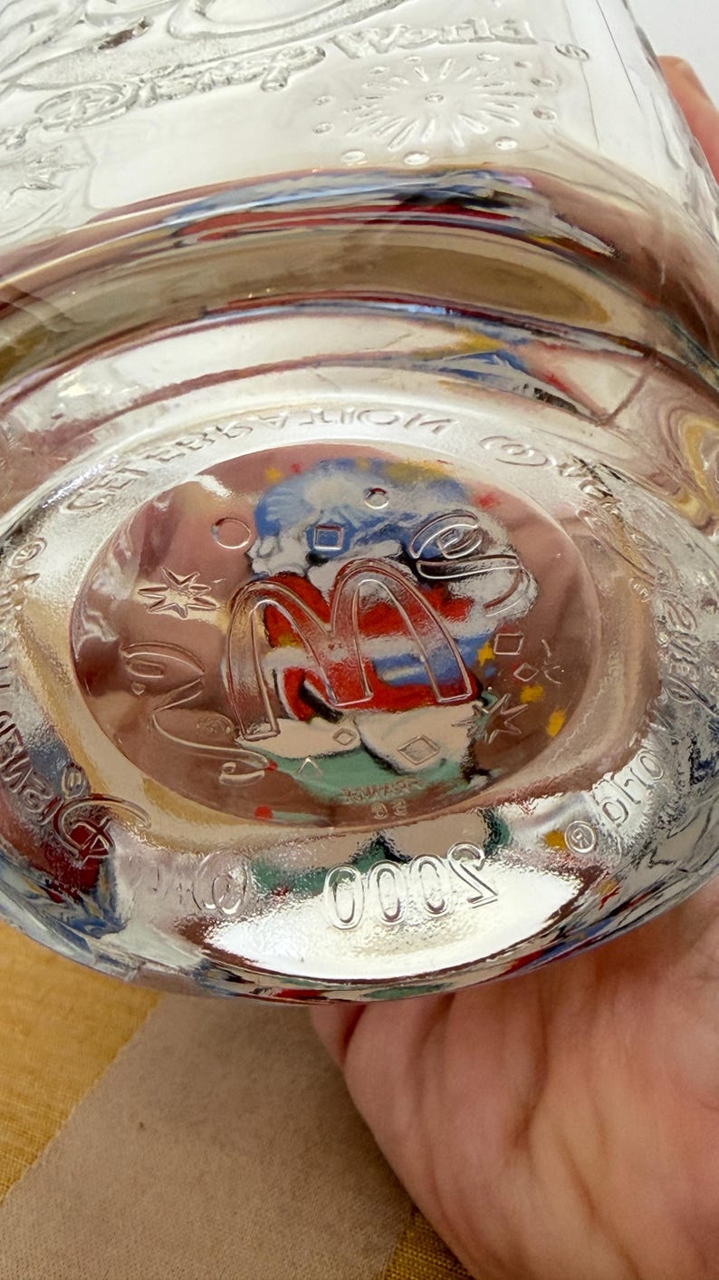 以下が含まれることがあります： 丸みを帯びた底を持つ透明なガラス瓶。底には、赤と黄色のマクドナルドのロゴがあり、カラフルな画像で囲まれています。「5000」の数字が底にエンボス加工されています。