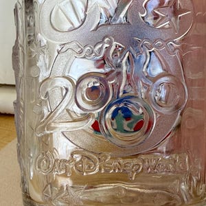 以下が含まれることがあります： エンボス加工が施された透明なガラスタンブラー。星や装飾的な要素とともに「25」の文字、「ウォルト・ディズニー・ワールド」と「セレブレーション」の文字が特徴です。グラスはクラシックな形状をしています。