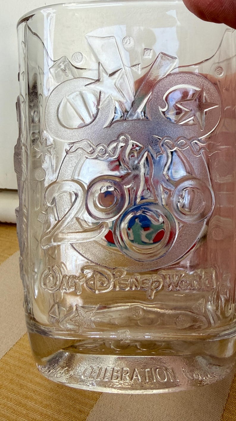 以下が含まれることがあります： エンボス加工が施された透明なガラスタンブラー。星や装飾的な要素とともに「25」の文字、「ウォルト・ディズニー・ワールド」と「セレブレーション」の文字が特徴です。グラスはクラシックな形状をしています。
