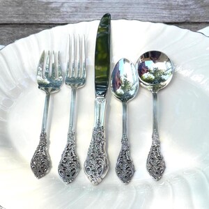 May include: A set of five ornate silver-colored utensils: two forks, a knife, a spoon, and a serving spoon. The utensils have detailed floral patterns on the handles and are arranged on a white, scalloped-edge plate. The knife blade reflects a green hue.