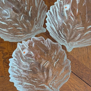 May include: Three clear glass leaf-shaped bowls. The bowls have a textured surface with leaf vein details. Each bowl has a scalloped edge. The bowls are on a wooden surface. The Blenko logo is visible on one of the bowls.