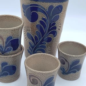 May include: A stoneware pitcher and matching tumblers. The set features a textured, tan background with blue and brown floral designs. The pitcher has a blue rim and the tumblers are stacked. The set is ideal for serving beverages.