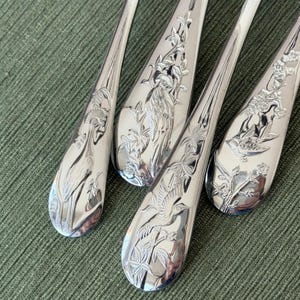 May include: Close-up of four silver spoons with detailed floral and bird engravings. The spoons have a shiny, reflective surface and are arranged diagonally. The intricate designs include various birds and botanical elements.